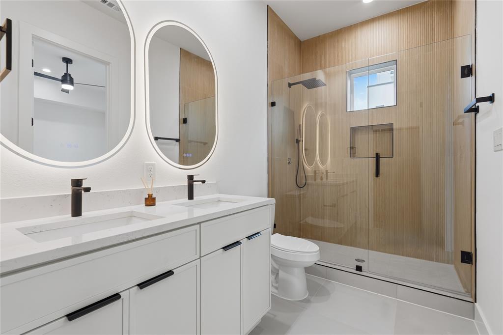 Double vanity featuring quartz countertops, matte black fixtures, illuminated oval mirrors, and a walk-in shower with wood-finish tile surround