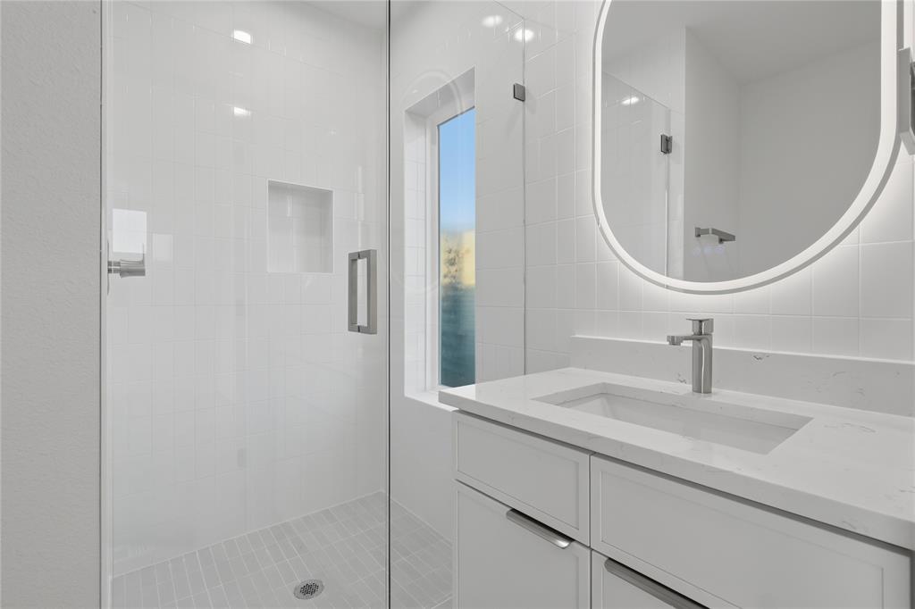 Bathroom featuring a white vanity with a stone countertop, an integrated rectangular sink, a frameless glass shower enclosure with a recessed tile niche, and an oval backlit mirror