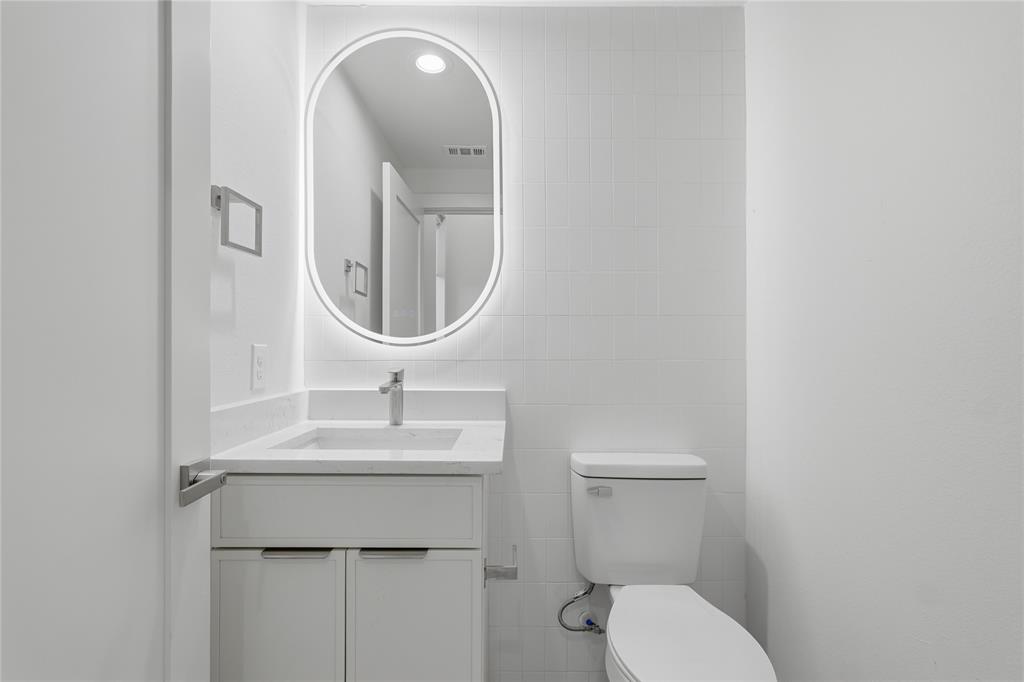 Bathroom featuring a white vanity with a composite countertop, integrated rectangular sink, wall-mounted oval LED mirror, and tiled wall surround