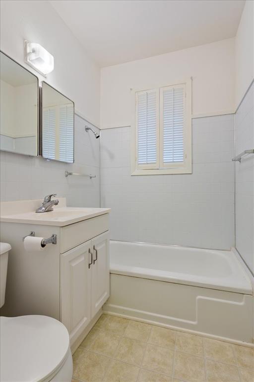 Bathroom featuring a vanity with a white cabinet and integrated sink, a tiled tub surround, a window with louvered shutters, wall-mounted lighting, and rectangular wall mirrors