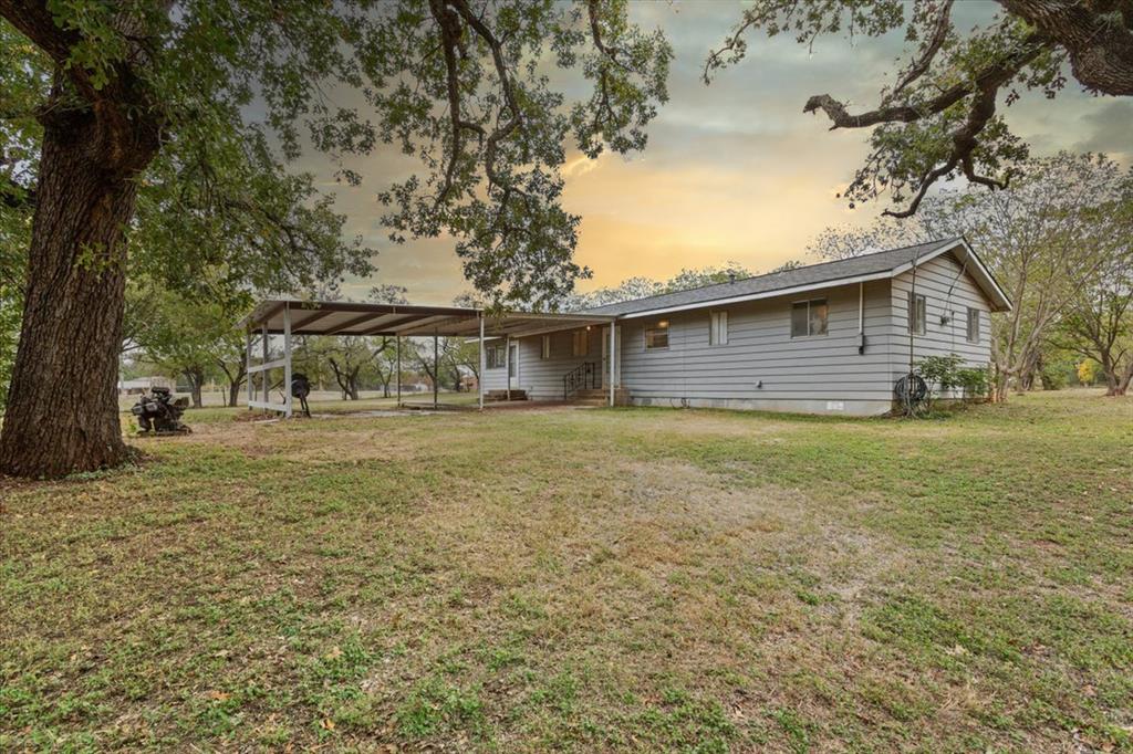 Single-story exterior featuring light-toned horizontal siding, a gabled roofline, an attached metal carport, and expansive grounds shaded by mature trees