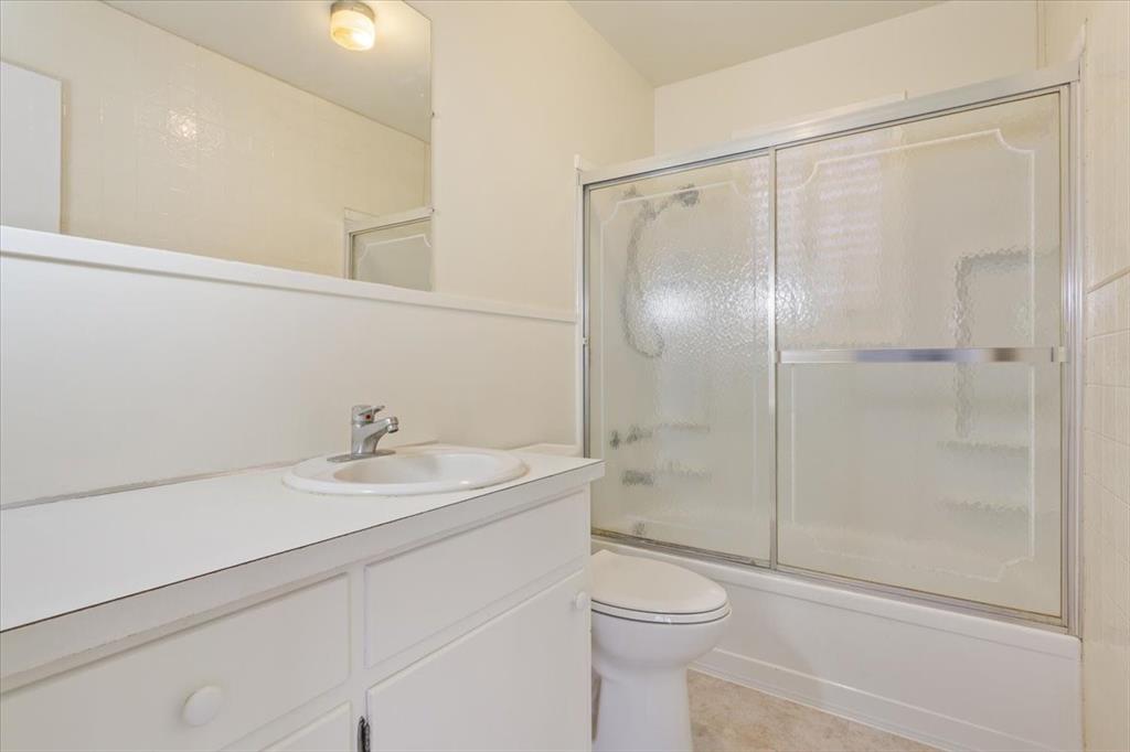 Bathroom featuring a white vanity cabinet with a solid surface countertop, integrated vanity sink, polished chrome faucet, and a shower-tub combination enclosed by sliding textured glass doors