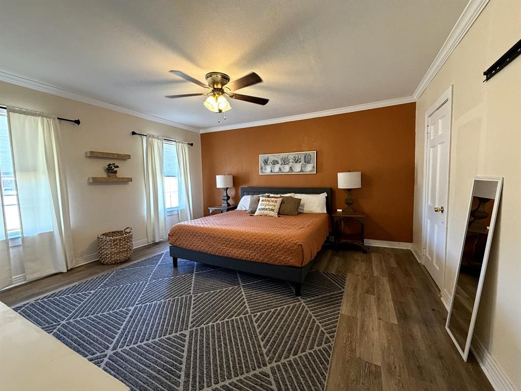 Room featuring wood-finish flooring, accent wall, ceiling fan with integrated lighting, crown molding, and wall-mounted shelving