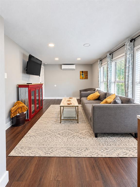 Living area featuring wood-finish flooring, recessed lighting, and a wall-mounted ductless climate control unit, characterized by large windows with horizontal blinds