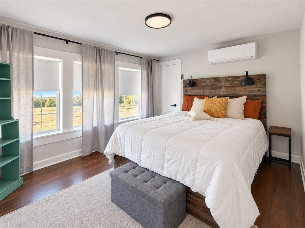 Wood-finish flooring, white paneled walls, multi-pane windows, wall-mounted heating and cooling unit, and flush-mount ceiling lighting