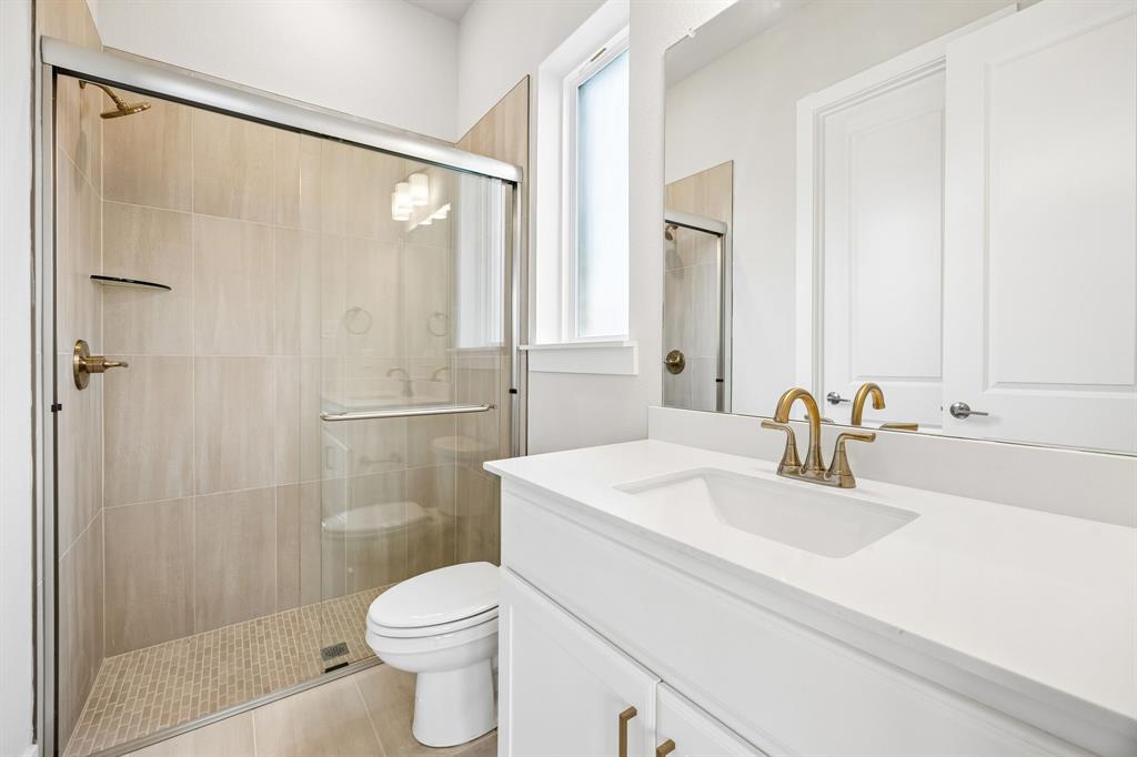 Bathroom featuring a white vanity with a rectangular sink, stone-finish countertop, brass-tone hardware, glass-enclosed shower with tiled walls and flooring, and a large wall mirror