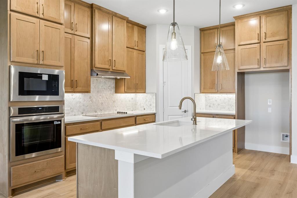 Kitchen featuring light wood-finish cabinetry, white stone countertops, herringbone-pattern tile backsplash, stainless steel wall-mounted oven and microwave, and a central island with pendant lighting