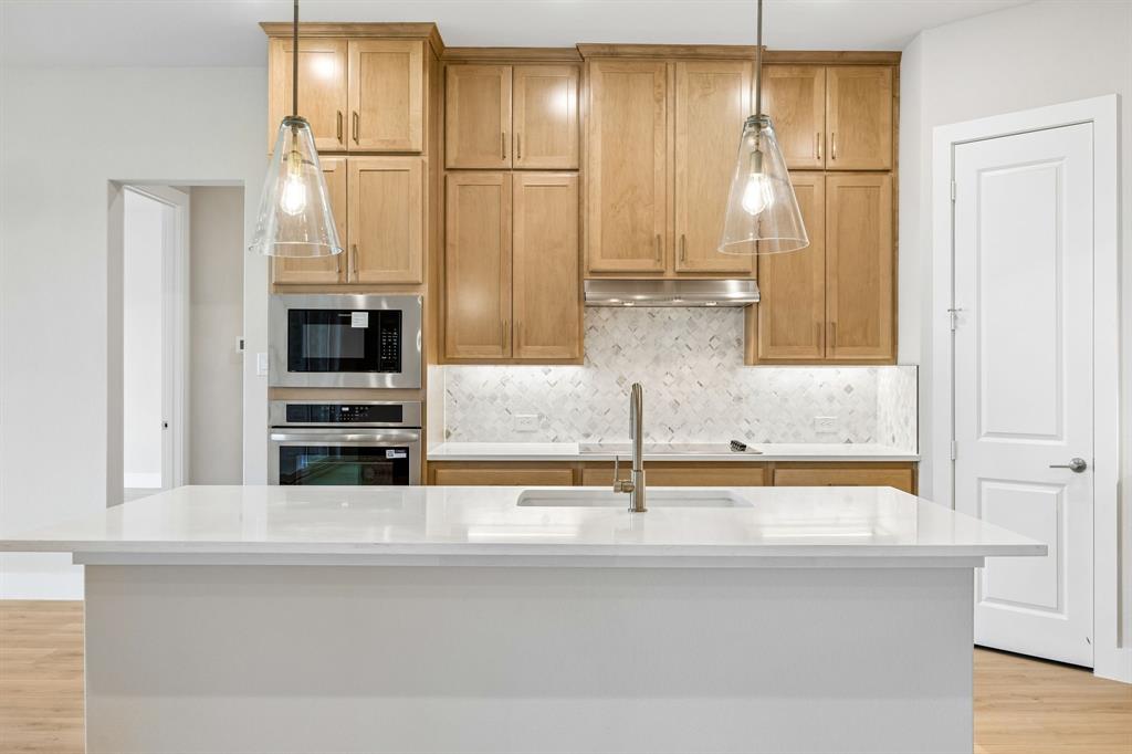 Kitchen featuring custom-height wood cabinetry, quartz countertop kitchen island with undermount sink, chevron-pattern tile backsplash, stainless steel built-in double wall oven and microwave, and glass pendant lighting