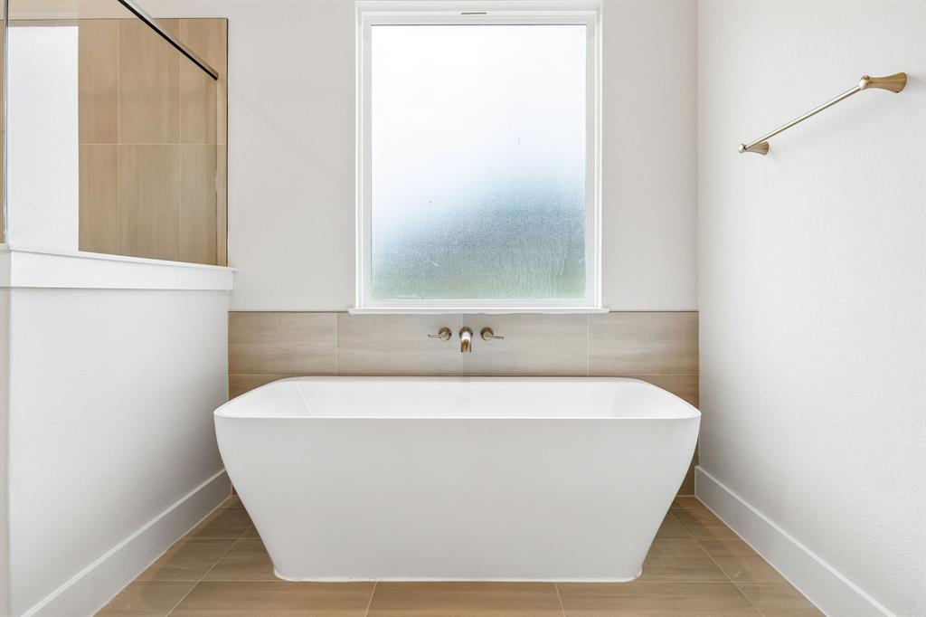 Freestanding soaking tub featuring a wall-mounted brushed-finish faucet, neutral-toned tile surround, tile flooring, and frosted window