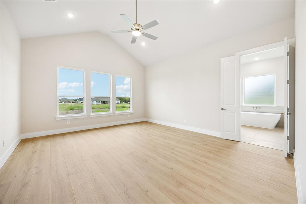 Vaulted ceiling featuring a central brushed-nickel finish ceiling fan, wood-finish flooring, three oversized windows, and double doors opening to a space with a freestanding soaking tub