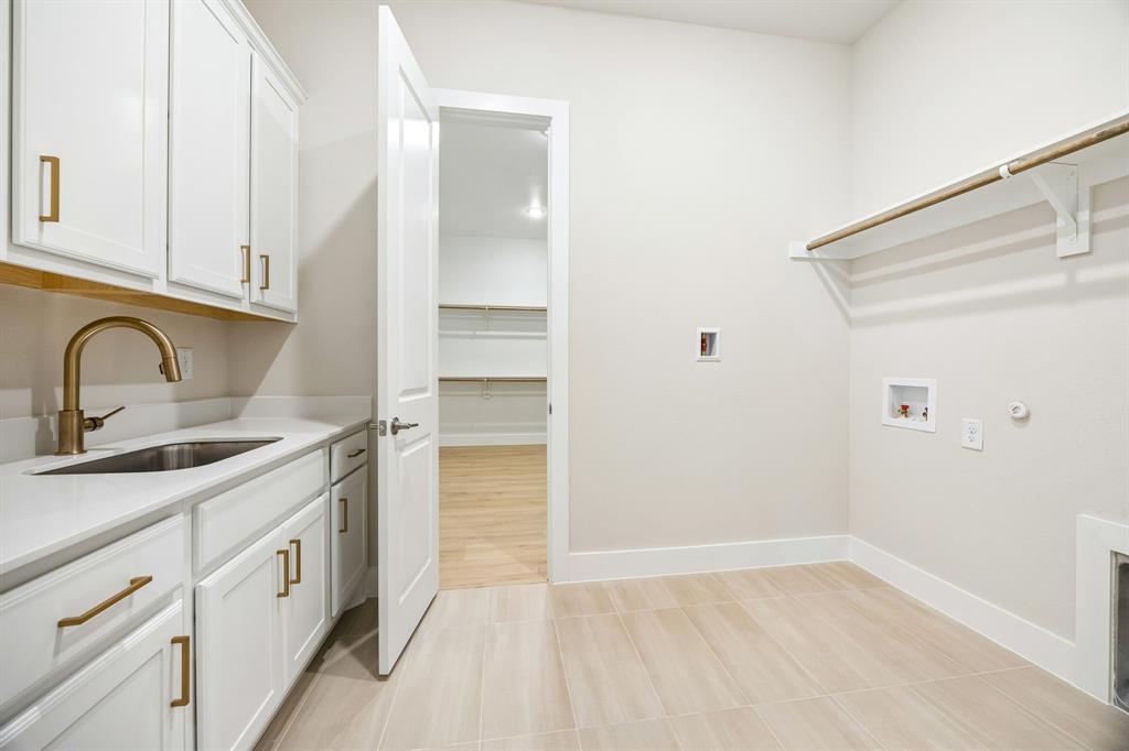 Laundry room featuring white cabinetry, a sink with brass-finish hardware, quartz countertops, tile flooring, and integrated wall shelving