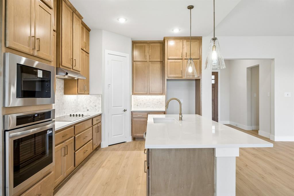 Kitchen featuring wood-finish cabinetry, quartz countertops, a central island with an undermount sink, built-in stainless steel double ovens, and glass pendant lighting