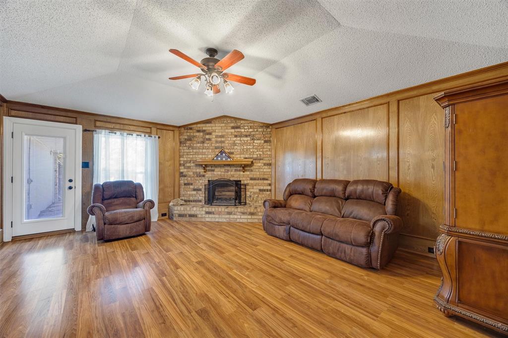 Vaulted ceiling with a central multi-blade ceiling fan, full-height wood paneling, wood-finish flooring, and a brick fireplace featuring a wood mantel