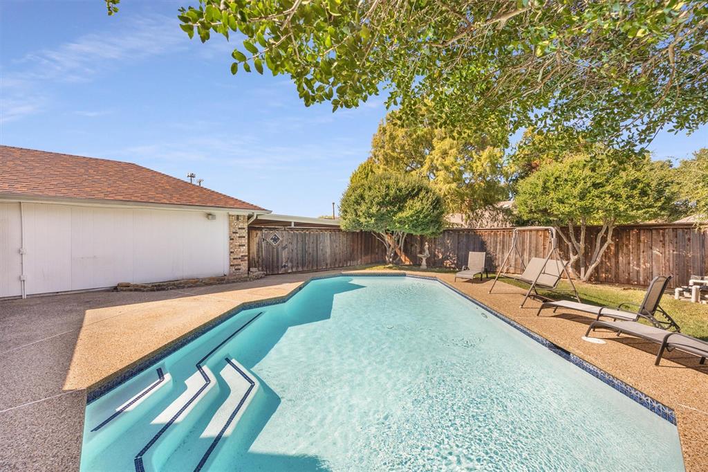 In-ground swimming pool with a stepped entry, surrounded by a textured aggregate pool deck, enclosed by a wood privacy fence