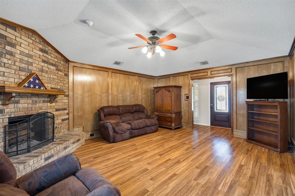 Vaulted ceiling with a central multi-blade ceiling fan, floor-to-ceiling brick fireplace with a wood mantel, wood-finish paneling, and wood-finish flooring