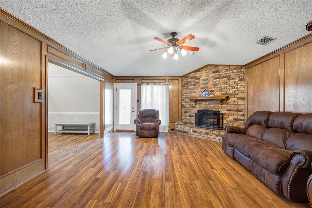 Living area featuring wood-finish flooring, full-height wood wall paneling, a vaulted ceiling with a central fan, and a masonry brick fireplace with a wood mantel
