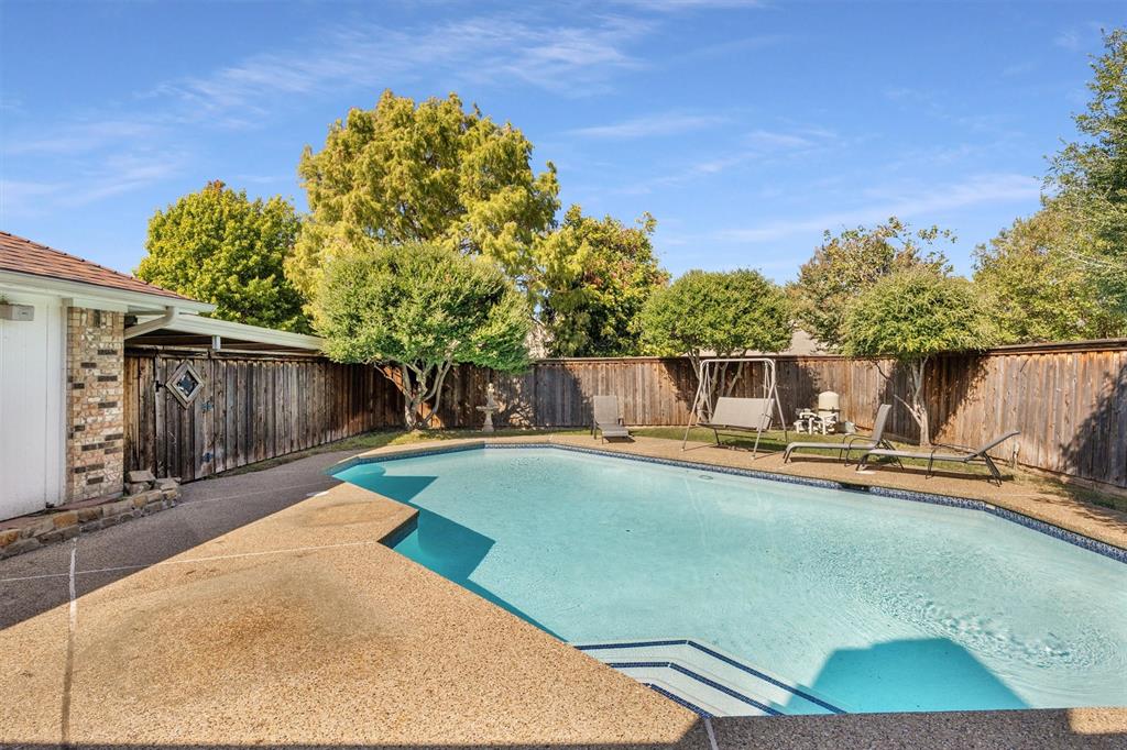 In-ground swimming pool featuring a textured concrete deck, tiered entry steps, privacy wood fencing, and mature landscaping