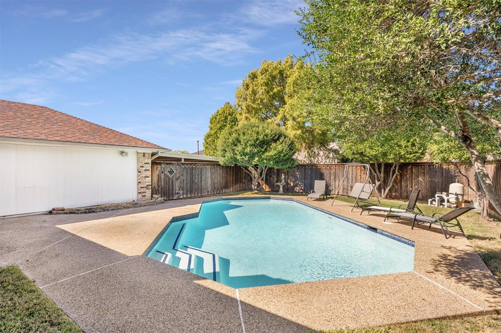 In-ground swimming pool featuring a multi-level entry, exposed aggregate pool decking, perimeter wood fencing, and mature trees