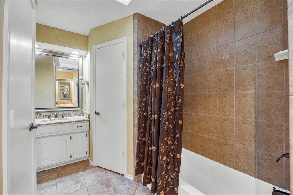 Bathroom featuring a stone-patterned vanity countertop, white cabinetry with integrated hardware, a framed wall mirror, tiled flooring, and a bathtub enclosure with decorative wall tile