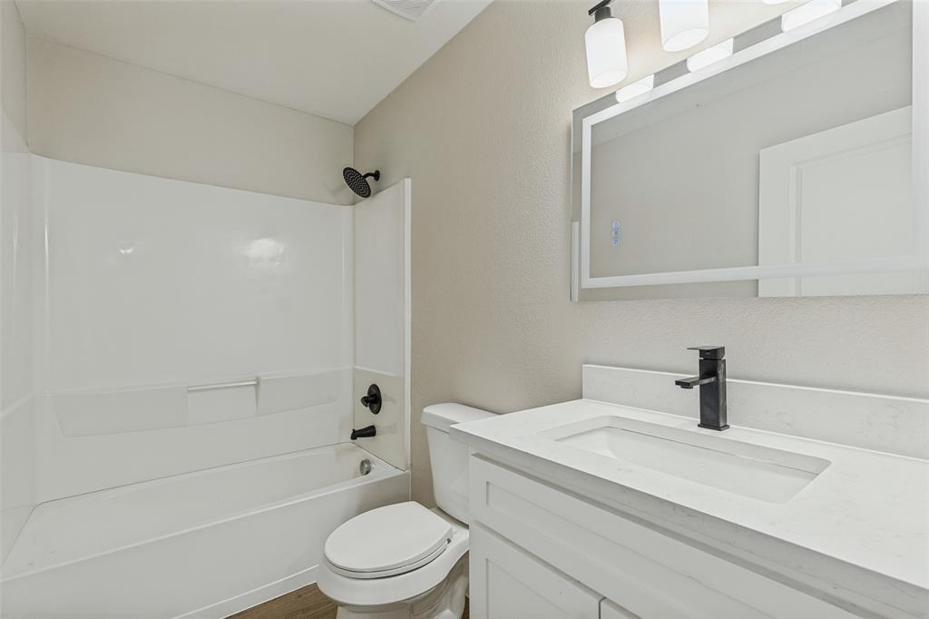 Bathroom featuring a stone-composite countertop, white shaker-style vanity cabinet, matte black faucet, integrated tub and shower surround with matte black fixtures, and a LED-backlit mirror