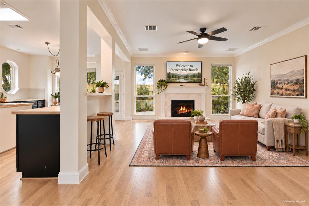 Open-concept room featuring light wood-finish flooring, a neutral-toned fireplace surround with a mantle, crown molding, a ceiling fan, and large windows