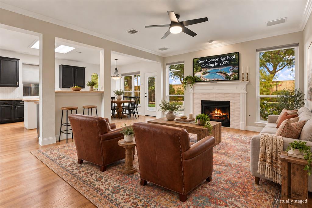 Living area featuring wood-finish flooring, fireplace with a mantle and tiled surround, crown molding, ceiling fan with integrated lighting, and multiple tall windows