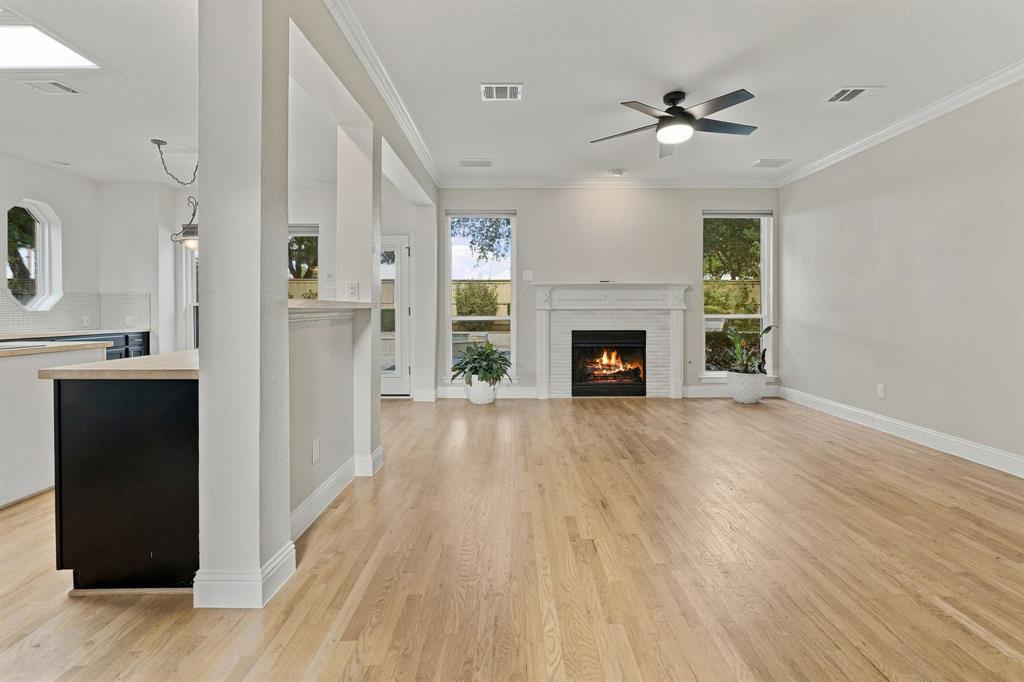 Living area featuring wood-finish flooring, a white brick fireplace with a decorative mantel, multiple floor-to-ceiling windows, and a ceiling fan with integrated lighting
