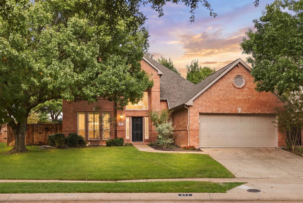 Traditional brick exterior featuring a multi-gabled roofline, an attached two-car garage, mature trees, and a front lawn