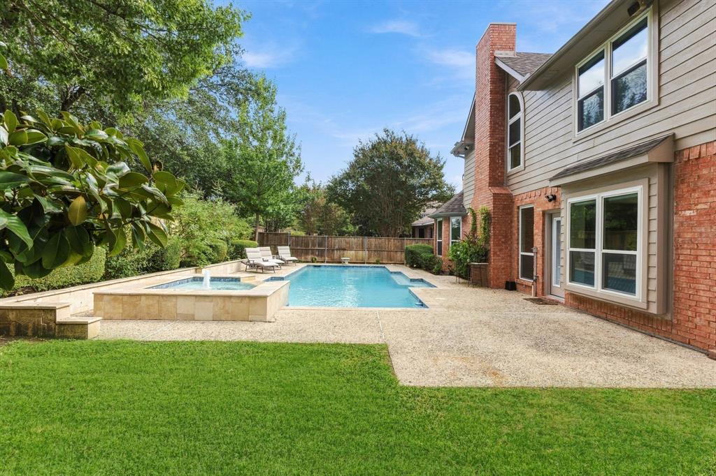 Rear exterior featuring a brick and siding facade, an in-ground swimming pool with an integrated spa, a tiered stone retaining wall, and a sprawling concrete patio