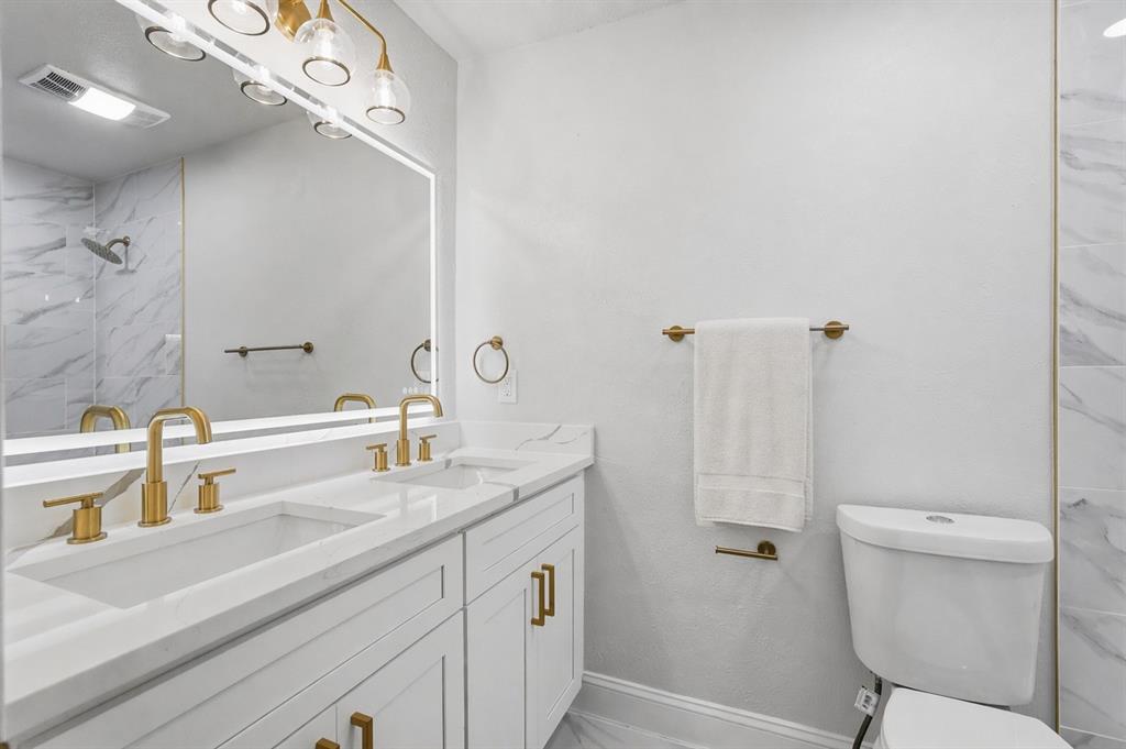 White cabinetry with stone-finish countertops, dual under-mount sinks, brass-toned fixtures, a full-width vanity mirror with integrated lighting, and tile-surround shower