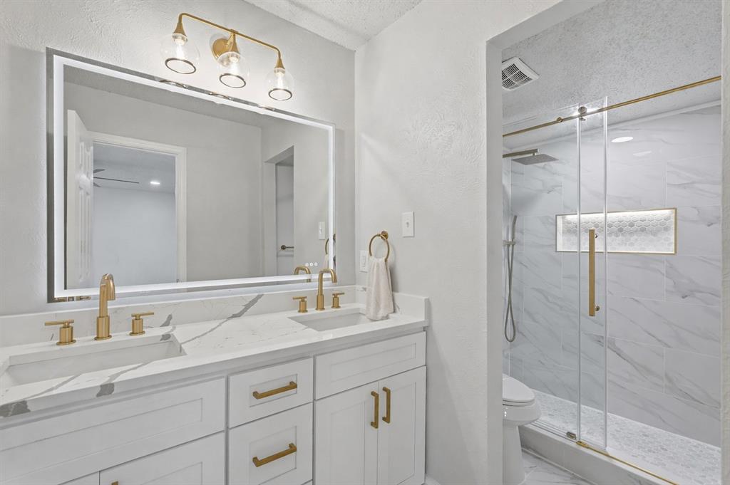 Dual-sink vanity with stone-surfaced countertop, integrated LED-backlit mirror, brass-finish hardware and fixtures, and a walk-in shower featuring a glass enclosure with a tiled built-in niche Dual-sink vanity with stone-surfaced countertop, integrated LED-backlit mirror, brass-finish hardware and fixtures, and a walk-in shower featuring a glass enclosure with a tiled built-in niche