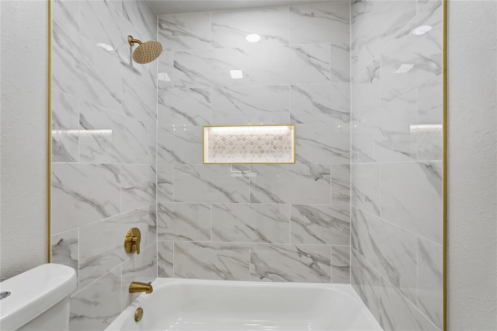 Bathtub with surround featuring large-format stone-patterned tile, gold-tone metal trim, integrated recessed niche with mosaic tiling, and gold-tone faucet and shower fixtures
