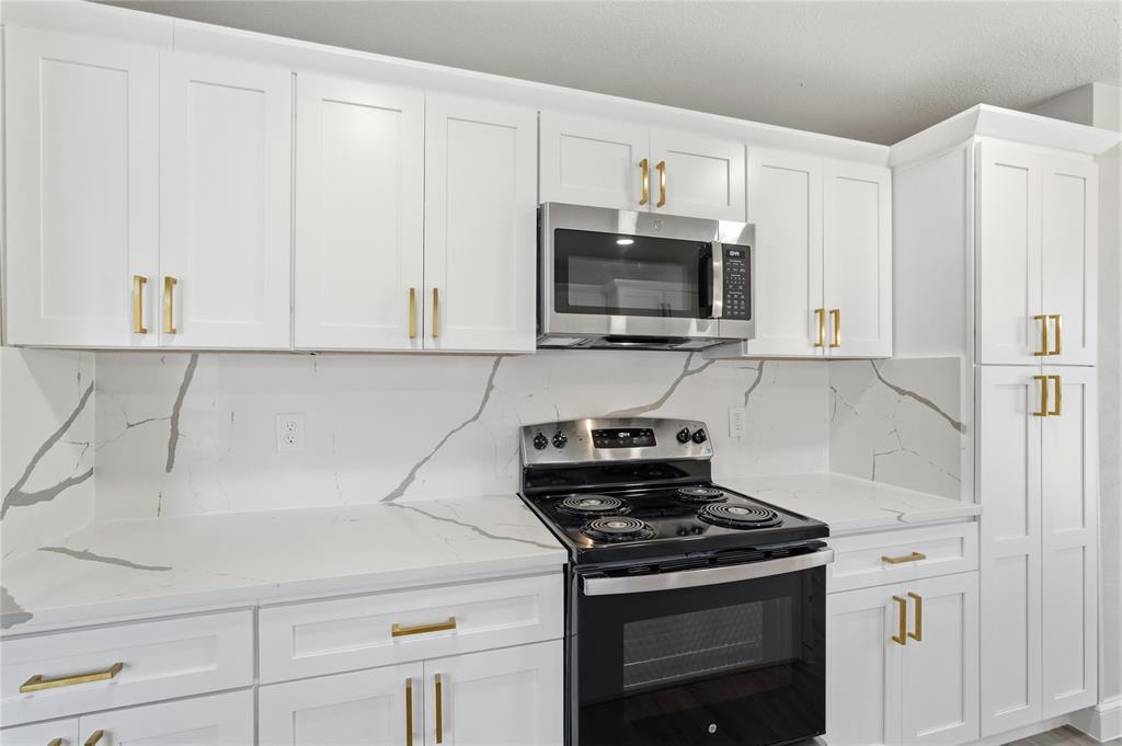 Shaker-style white cabinetry with brushed gold hardware, stone-finish countertops, full-height stone-finish backsplash, and stainless steel appliances