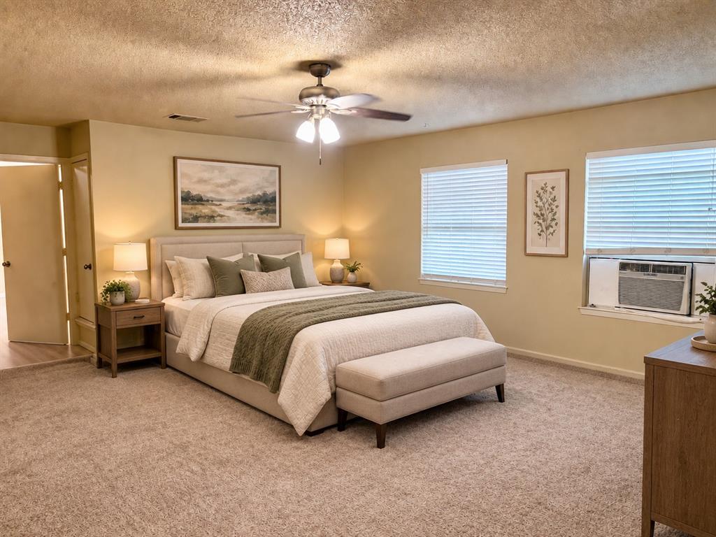 Spacious room featuring neutral carpeting, a central ceiling fan with integrated lighting, dual windows with horizontal blinds, and a built-in wall air conditioning unit