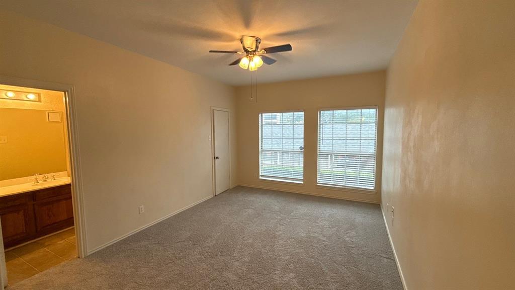 Neutral-toned interior featuring wall-to-wall carpeting, dual windows with integrated blinds, a ceiling fan with multi-light fixture, and a built-in wood cabinet vanity