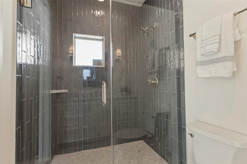 Walk-in shower featuring vertical subway tile surround, frameless glass door, wall-mounted rainfall showerhead, and pebble-style stone flooring