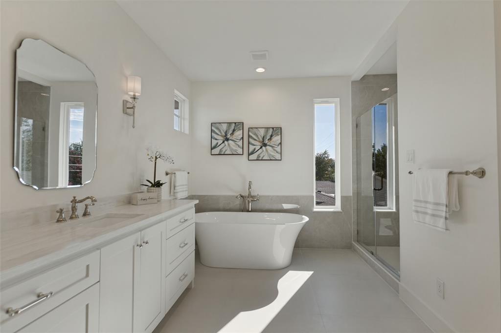 Freestanding soaking tub with floor-mounted faucet, glass-enclosed shower, white vanity cabinet with stone-finish countertop, scalloped-edge wall mirror, and dual vertical windows
