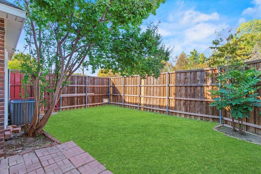 Fenced backyard featuring a manicured lawn, brick patio, mature trees, and wooden privacy fencing