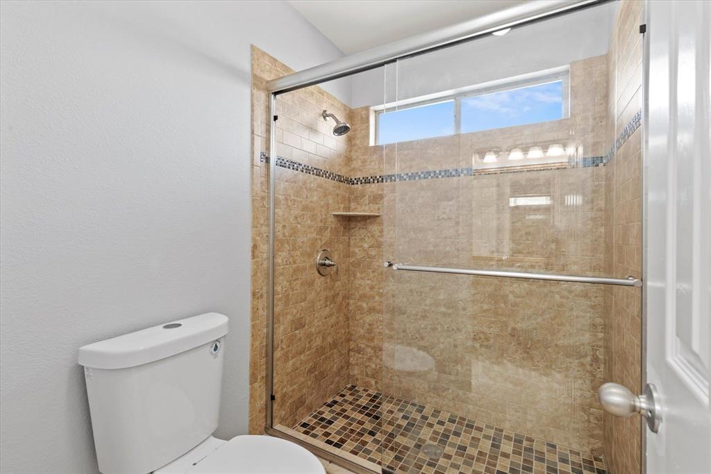 Upstairs Jack and Jill Bathroom.Walk-in shower featuring a sliding glass enclosure, stone-finish tile surround, decorative mosaic tile inlay, integrated shelving, and a horizontal transom window