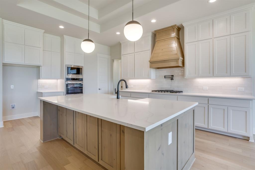 Kitchen featuring white shaker cabinetry, a central island with wood-finish base and light-toned countertop, a custom wood vent hood, globe pendant lighting, and built-in double wall ovens