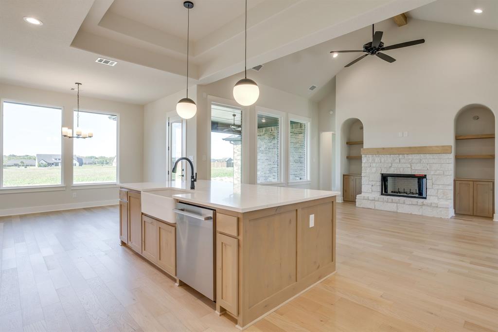 Open-concept floor plan featuring light wood-finish flooring, a central kitchen island with a farmhouse sink and quartz countertops, vaulted ceilings, and a floor-to-ceiling stone fireplace flanked by built-in shelving