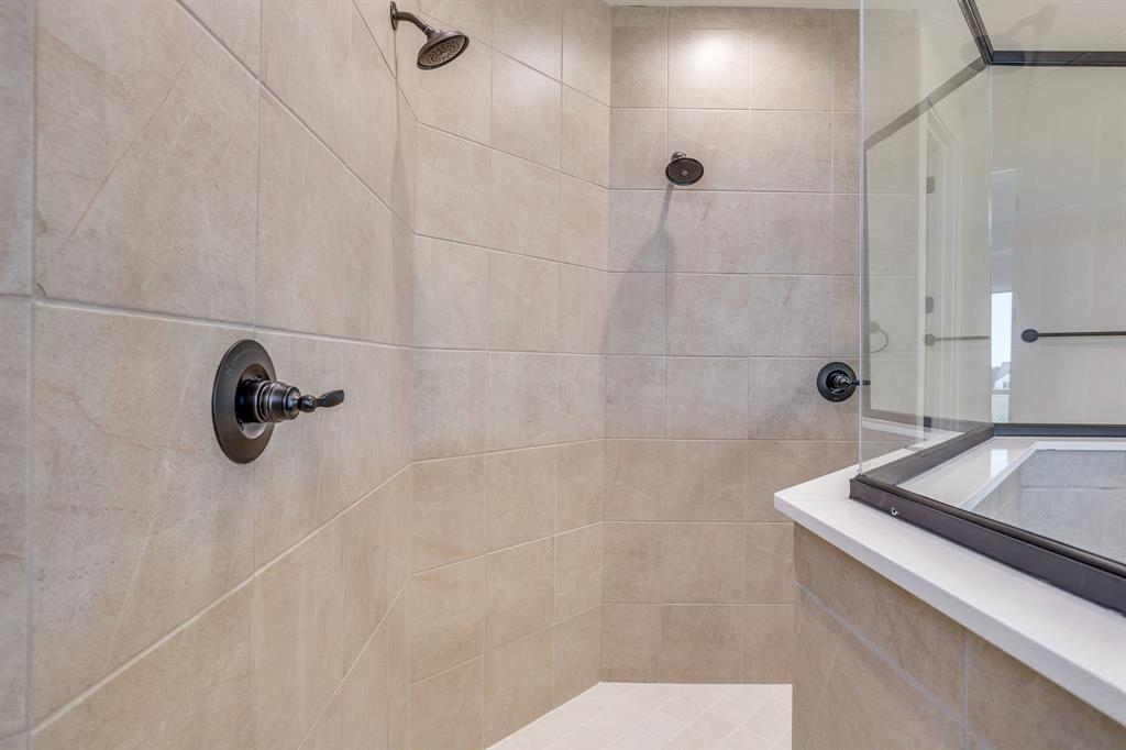 Walk-in shower featuring large-format neutral tile, dual wall-mounted shower heads, dark-finish hardware, and glass enclosure with a solid-surface threshold