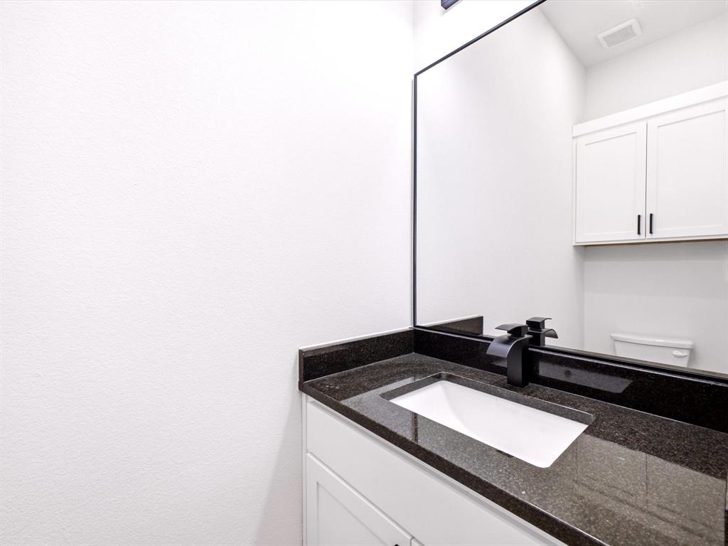 Vanity featuring a dark stone countertop, integrated rectangular sink, matte black faucet, full-width mirror, and white shaker-style cabinetry with overhead storage