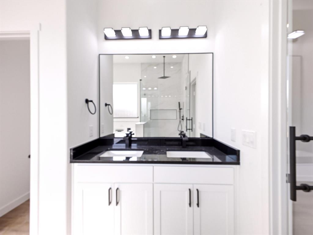 Double vanity featuring white shaker-style cabinetry, dark countertop with dual rectangular undermount sinks, matte black wall-mounted fixtures, and a large frameless mirror with dual modern vanity lights
