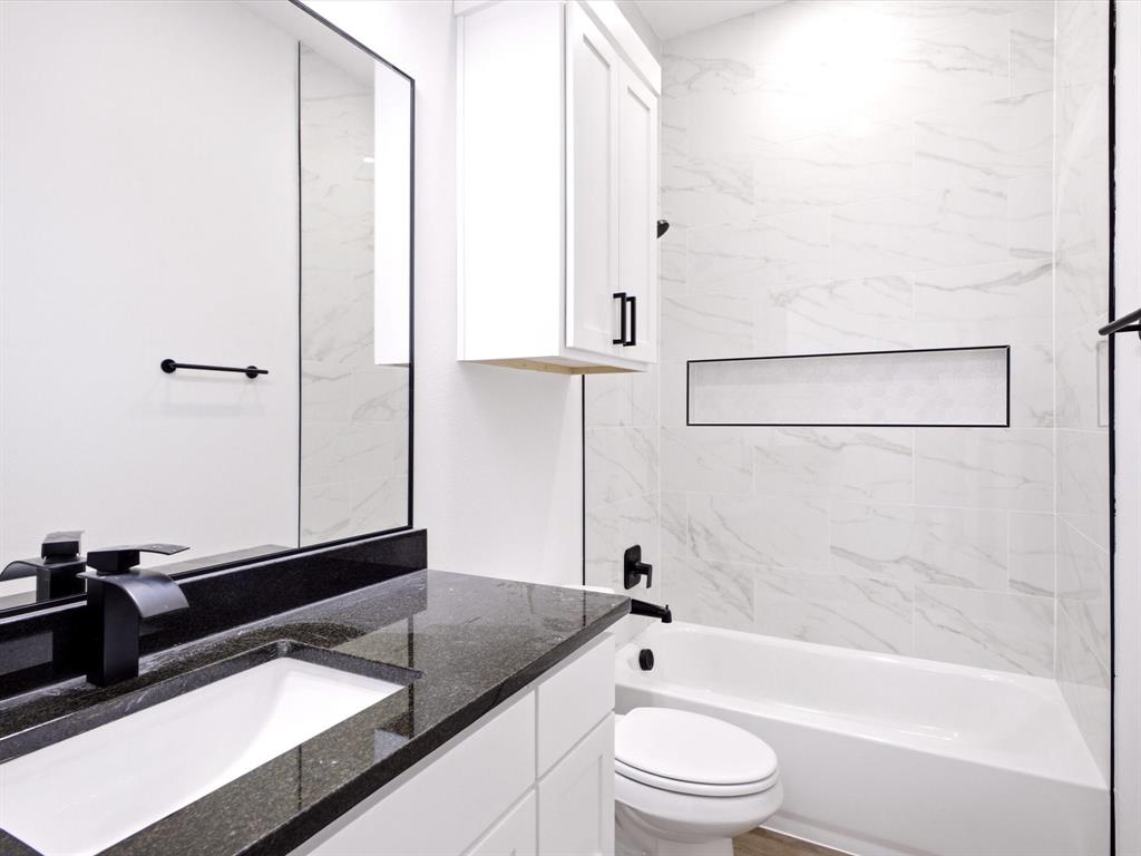 White vanity with dark stone-finish countertop and undermount sink, complemented by a matte black faucet, wall-mounted cabinet, and shower area featuring large-format stone-finish tile surround with integrated recessed shelving