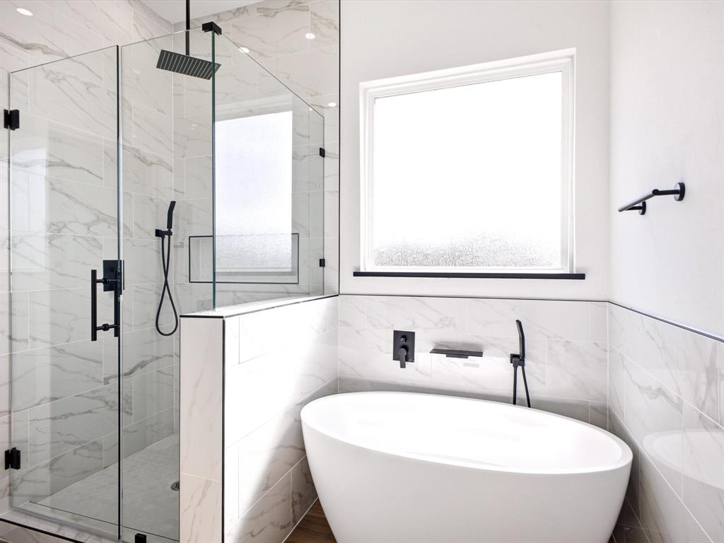 Walk-in glass shower featuring matte black hardware, rainfall showerhead, and wall-mounted hand sprayer, paired with a freestanding soaking tub, matte black wall-mounted faucet, and floor-to-ceiling stone-look tile