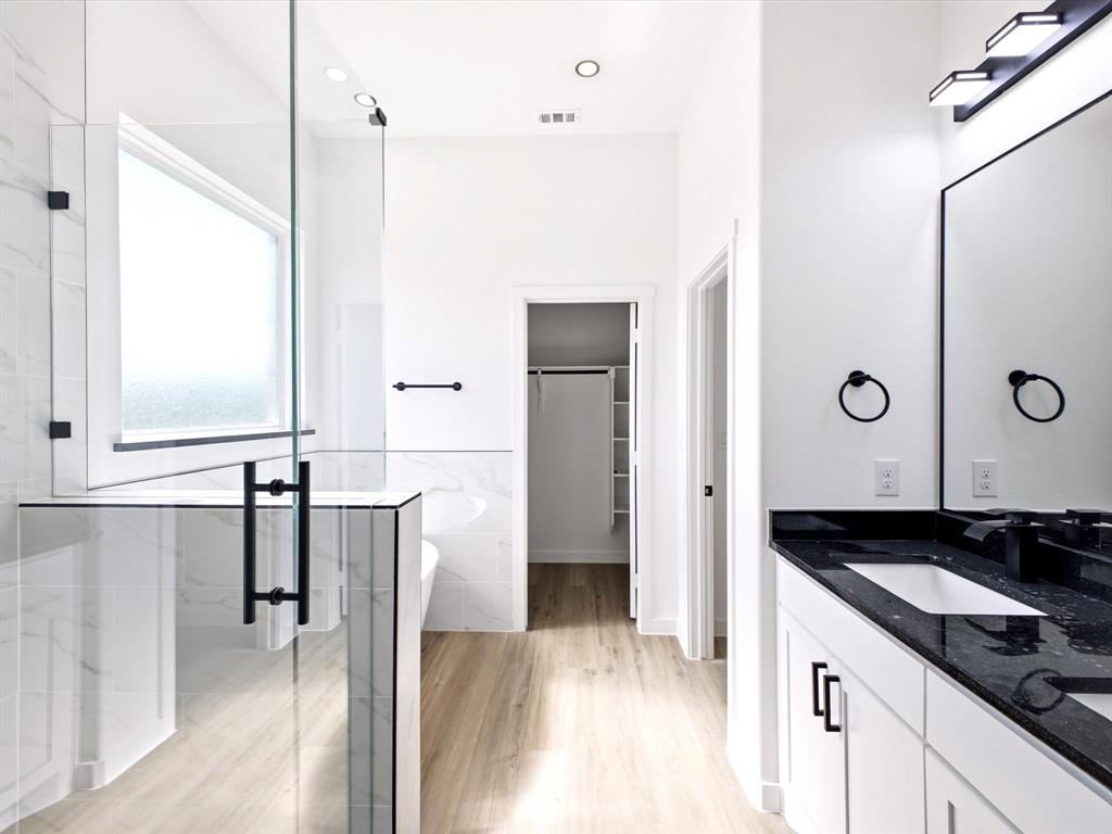 Bathroom featuring a frameless glass shower enclosure, stone countertop vanity, matte black fixtures, wood-finish flooring, and built-in closet shelving