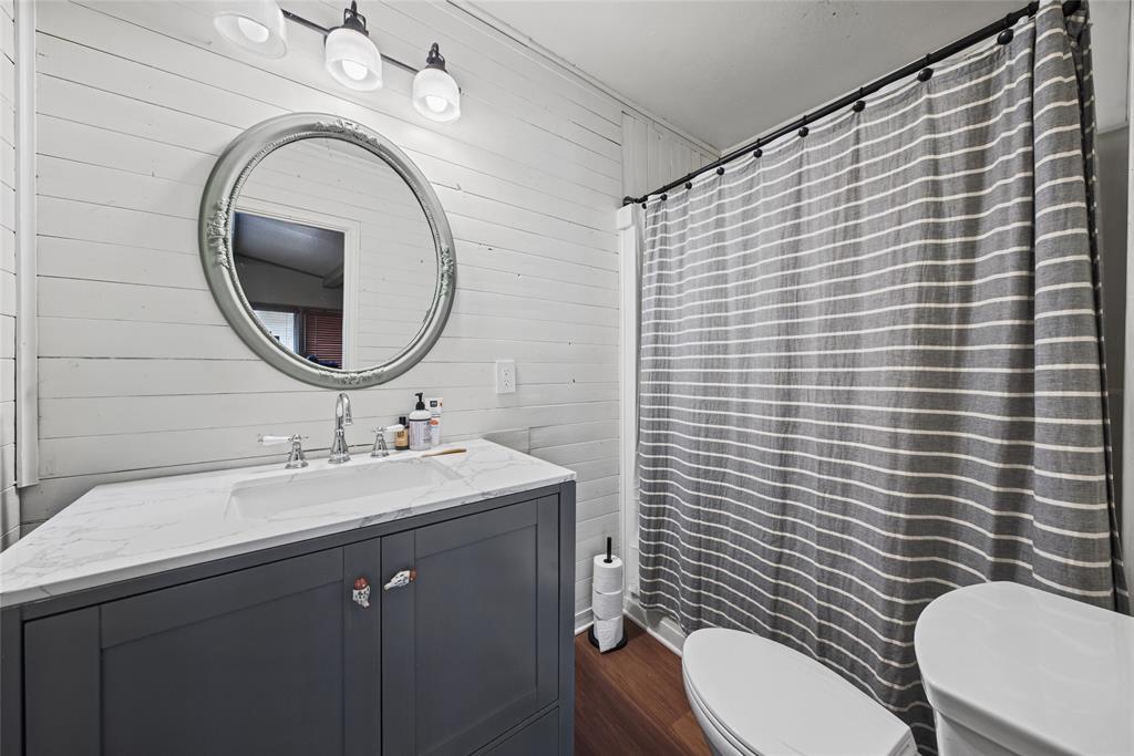 Dark gray vanity with a stone-top integrated sink, horizontal shiplap wall paneling, circular mirror with decorative frame, wood-finish flooring, and a three-light vanity fixture