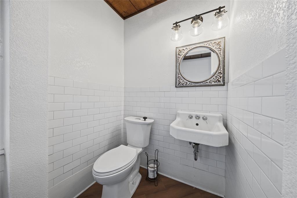 Wall-mounted porcelain sink, white subway tile wainscoting, wood-finish flooring, and three-light vanity fixture