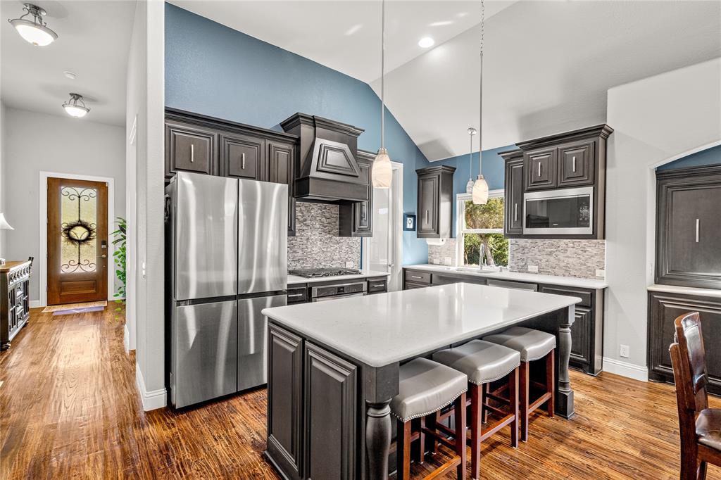 Kitchen featuring dark-stained cabinetry, a central island with a stone-composite countertop, stainless steel refrigerator, patterned tile backsplash, and vaulted ceilings